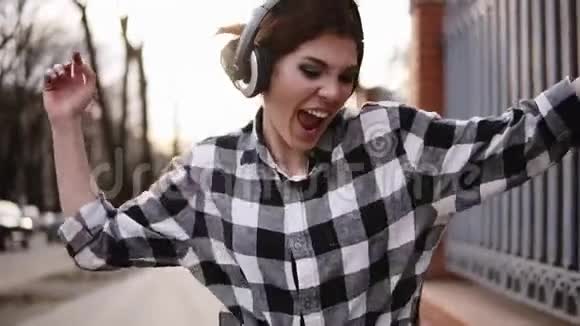 活泼微笑的女孩穿着衬衫走在街上跳舞有趣的时髦风格耳机表达式视频的预览图