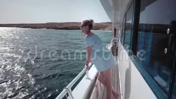 一位年轻漂亮的高加索女人穿着蓝色连衣裙和太阳镜在游艇上的肖像看着一个荒芜的人视频的预览图