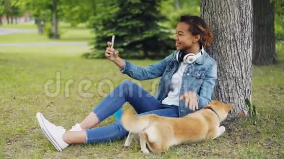 漂亮的年轻女子非裔美国学生正在用智能手机打视频电话并在草坪上抚摸她的狗视频的预览图