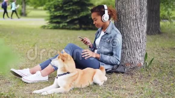 漂亮的非洲裔美国妇女正在用耳机听音乐并使用智能手机坐在公园的草地上视频的预览图
