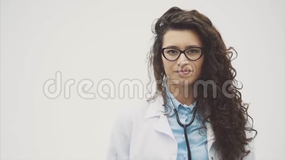 年轻的医生女孩穿着白色的晨衣展示一个手势课站在灰色背景上视频的预览图