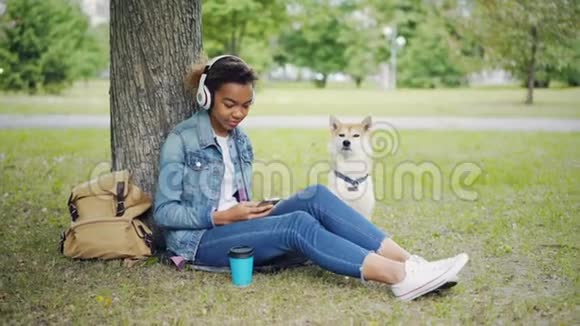 现代非洲裔美国女孩正在用耳机听音乐并使用智能手机在城市公园与宠物放松视频的预览图