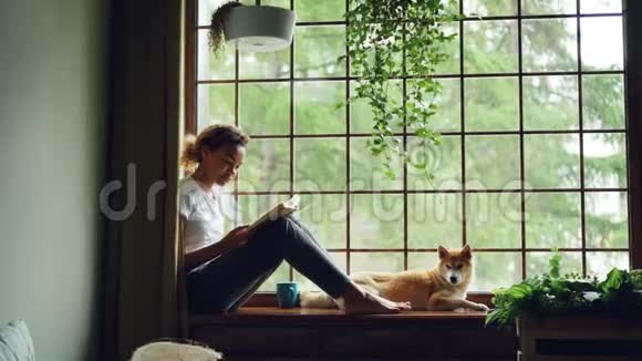 一个穿着休闲服的漂亮女孩赤脚坐在窗台上看书旁边躺着一只可爱的宠物狗视频的预览图