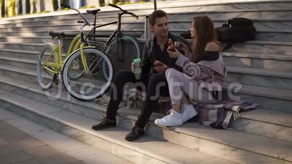 美丽的情侣坐在楼梯上吃牛角面包在户外喝咖啡坐在格子呢上视频的预览图