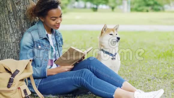 善良的混血女人正在公园看书一起抚摸她坐在树下草坪上的狗智能智能视频的预览图