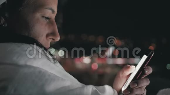 女孩在一条毯子里晚上在模糊的城市灯光前使用智能手机视频的预览图