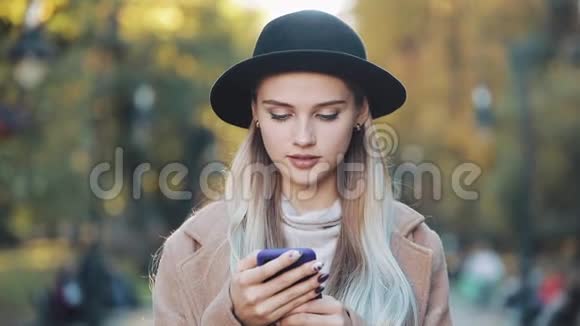 时尚年轻的女人用智能手机站在秋天的公园里看着相机她穿着外套视频的预览图