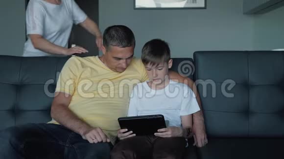父亲母亲和儿子坐在沙发上看电影在平板电脑上玩游戏一个幸福的家庭视频的预览图