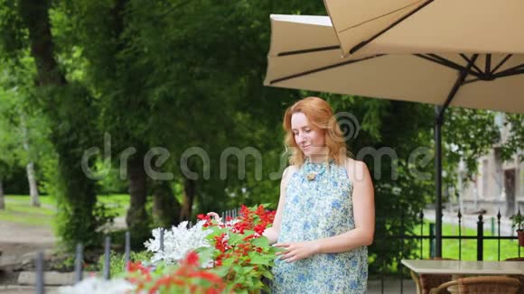 一个迷人的女人站在夏天的咖啡馆里抚摸着鲜花看着相机金发女人站在视频的预览图