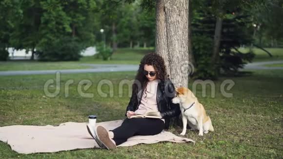 放松的女孩正在看书在公园的草坪上喝着外卖咖啡而她可爱的狗闻着地面视频的预览图