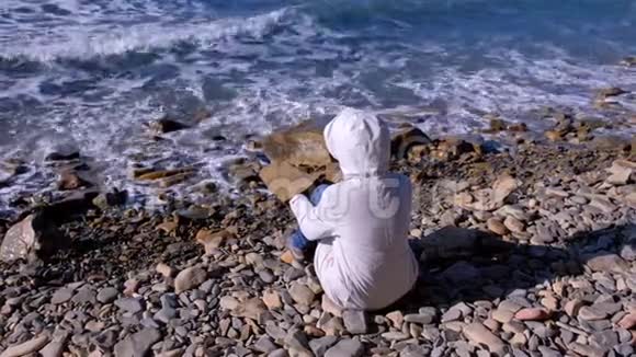 穿着白色羽绒服和风帽的女人坐在岸边看着汹涌的海浪拍打着石头视频的预览图