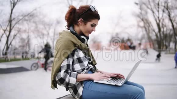 一位年轻时髦的黑发女郎正坐在长凳上膝盖上放着一台笔记本电脑打字戴着耳机戴着眼镜在视频的预览图