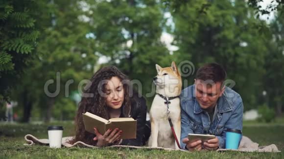 幸福的夫妻在公园里休息男人在用智能手机而女人在看书他们的狗坐在他们之间视频的预览图
