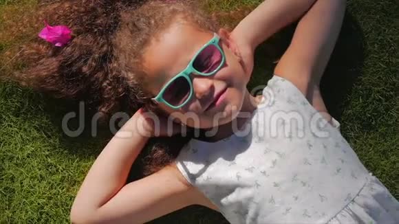 一个可爱的孩子的肖像一个穿着白色衣服和粉色眼镜的美丽的小女孩躺在草地上视频的预览图
