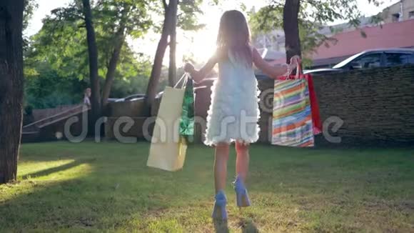 新买的时髦的小女孩穿着高跟鞋手里拿着很多购物袋背着背光走在草地上视频的预览图