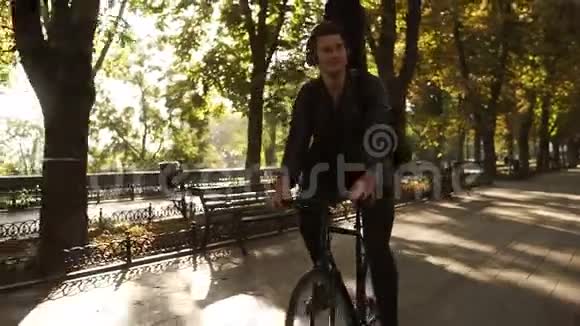 快乐微笑的年轻人穿着黑色便服戴着耳机骑着自行车骑在铺好的城市公园旁黑色吉他演奏视频的预览图