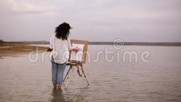 这位穿着蓝色牛仔裤和白色衬衫的女艺术家站在水里画架直到脚踝为她的未来工作视频的预览图