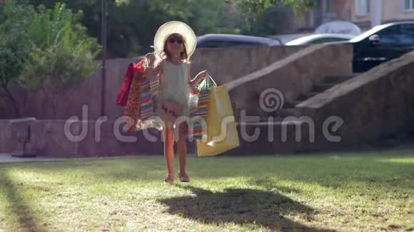 购物的孩子穿着时髦的衣服在参观商店后手里拿着很多袋子走在草坪上视频的预览图