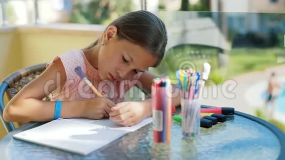 可爱的女孩在笔记本上画一支铅笔坐在阳台上的玻璃桌旁视频的预览图
