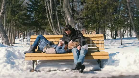 女人睡觉男人看电话男人和女人一起在冬季城市公园的长凳上休息冬日阳光明媚视频的预览图