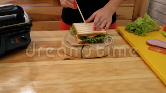 厨房里的女孩把一根木棍插入刚组装好的三明治里夹着新鲜绿叶莴苣的白色面包三明治视频的预览图