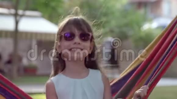 快乐的童年快乐的小女孩戴上太阳镜在吊床上摆动在大自然中微笑特写视频的预览图