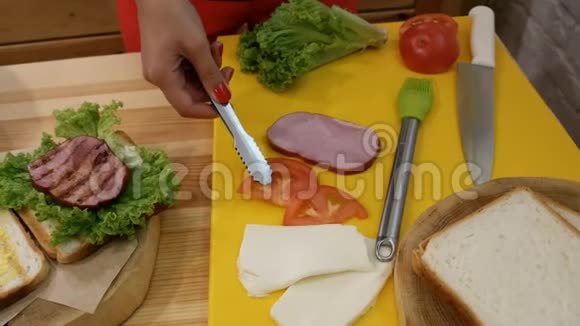在厨房里做三明治一只带钳子的雌性手拿着一个新鲜的番茄切片用一片生菜放在面包上视频的预览图