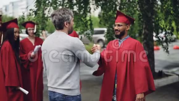 骄傲的老师正在祝贺学生在毕业的那一天他握手拥抱他站在校园里视频的预览图