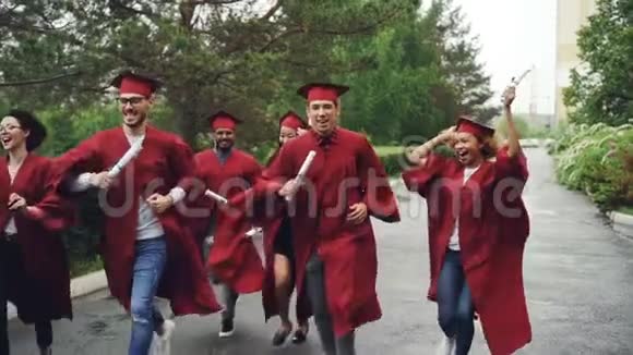 欢快的毕业生在雨中缓慢地奔跑挥舞着文凭穿着红色的长袍和迫击炮笑着视频的预览图
