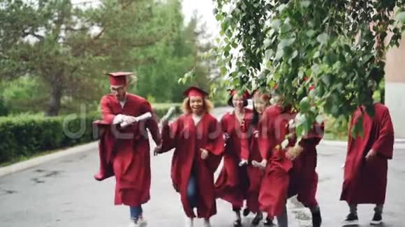 快乐的毕业生在校园里缓慢地奔跑挥舞着文凭微笑着穿着红色的长袍和帽子美丽的树视频的预览图