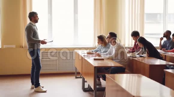 穿着便服的男教授正在和一群坐在教室里的桌子旁做笔记的学生交谈大型视频的预览图