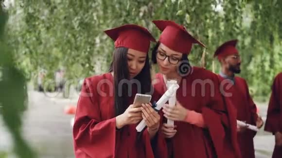女毕业生们用智能手机看屏幕有说有笑站在户外拿着文凭女孩们视频的预览图