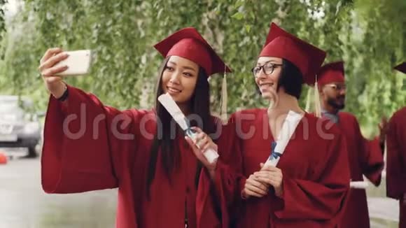 漂亮的女毕业生们正在用智能手机用毕业证书卷轴自拍女人们在摆姿势和微笑视频的预览图