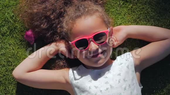 一个可爱的孩子的肖像一个美丽的小女孩穿着白色的连衣裙和粉红色的眼镜躺在草地上视频的预览图
