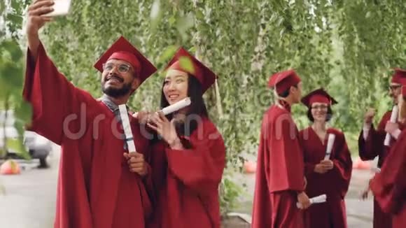 两名混血学生正拿着毕业证书带着灰泥板和礼服自拍盖伊正拿着视频的预览图