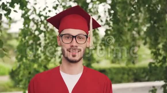 戴着眼镜毕业礼服和灰泥板的留着胡子的青年毕业学生的肖像微笑着看着视频的预览图