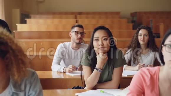 漂亮的亚洲女孩在大学演讲时举手提问她的同学坐在课桌前视频的预览图