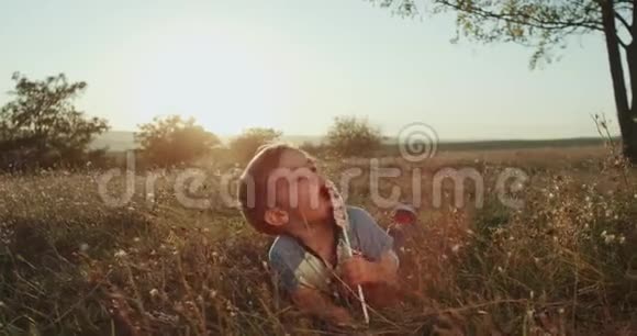 可爱的小男孩在大自然美丽的夕阳下吃着一根棒棒糖躺在草地上视频的预览图