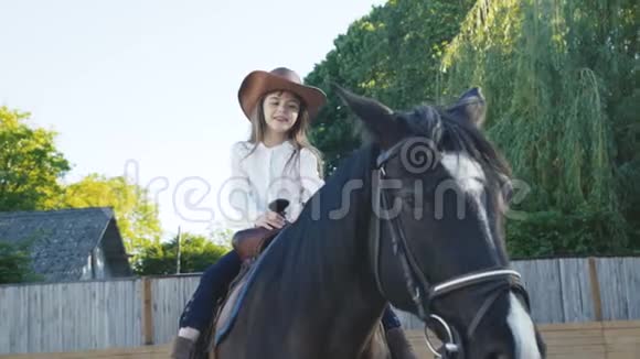 戴帽子的漂亮小女孩骑着一匹黑色的母马在竞技场上4K视频的预览图