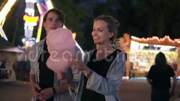 年轻的时髦夫妇晚上正在游乐园玩以粉色棉花糖的形式互相喂养视频的预览图