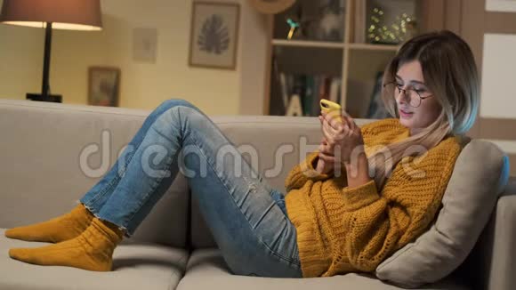 年轻女子躺在客厅的沙发上用智能手机发短信微笑的金发女孩在看留言视频的预览图