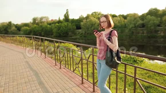 欧洲城市使用智能手机戴眼镜的时髦女孩她站在桥上的户外日落视频的预览图