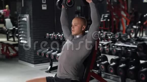 强壮的肌肉男正在黑暗的健身房训练运动员坐在长凳上按哑铃训练肩膀和视频的预览图