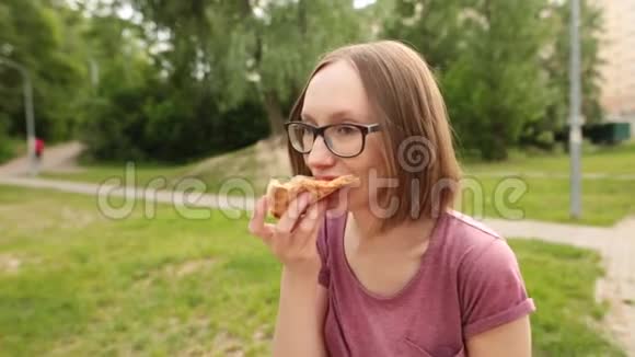 时髦的高加索女人戴着眼镜坐在长凳上吃着一片美味的披萨视频的预览图