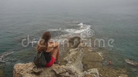 女游客背着背包坐在晴朗的天空和平静的海洋背景上的岩石上视频的预览图
