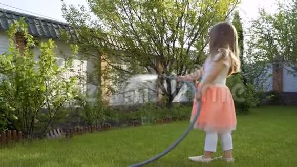一个漂亮的小女孩穿着连衣裙用水管浇草和灌木丛视频的预览图