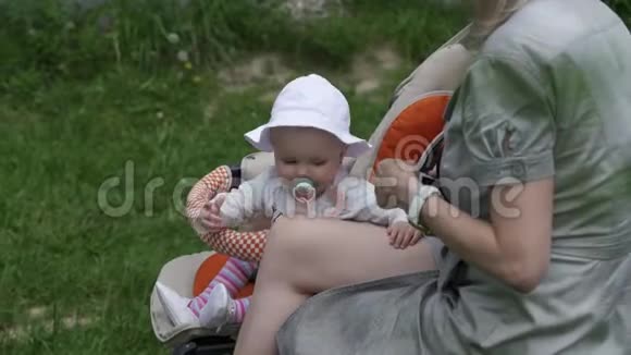 妈妈带着一个小女儿在公园里的长凳上休息一岁的孩子坐在马车里玩视频的预览图