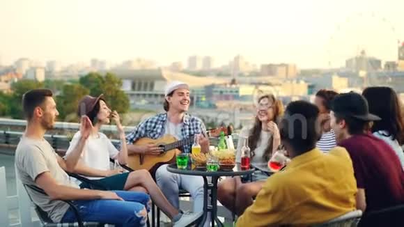 快乐的音乐家正在和他的朋友们一起弹吉他坐在屋顶的桌子上唱歌和笑娱乐视频的预览图