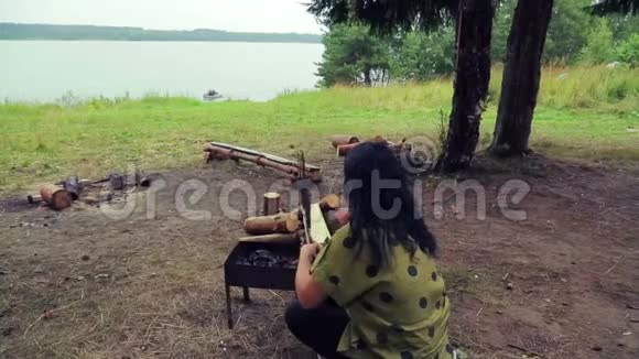 河岸上的一个女人坐在烧烤处用一条鱼把烤架翻过来视频的预览图