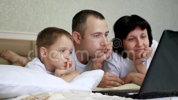快乐的爸爸妈妈和孩子都躺在床上看着笔记本电脑屏幕慢悠悠地笑着视频的预览图